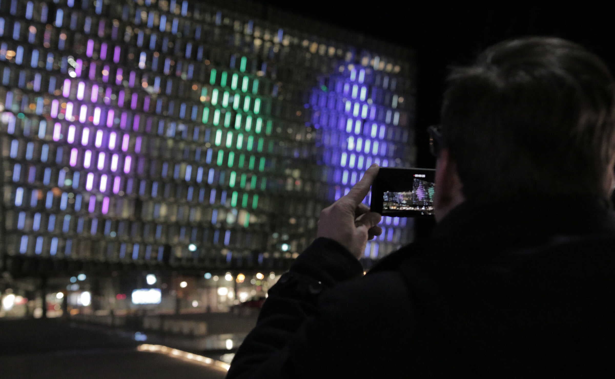 Festival attendee filming the Dice projection on Harpa's façade with a smartphone, with three colored sequencer columns visible across the building